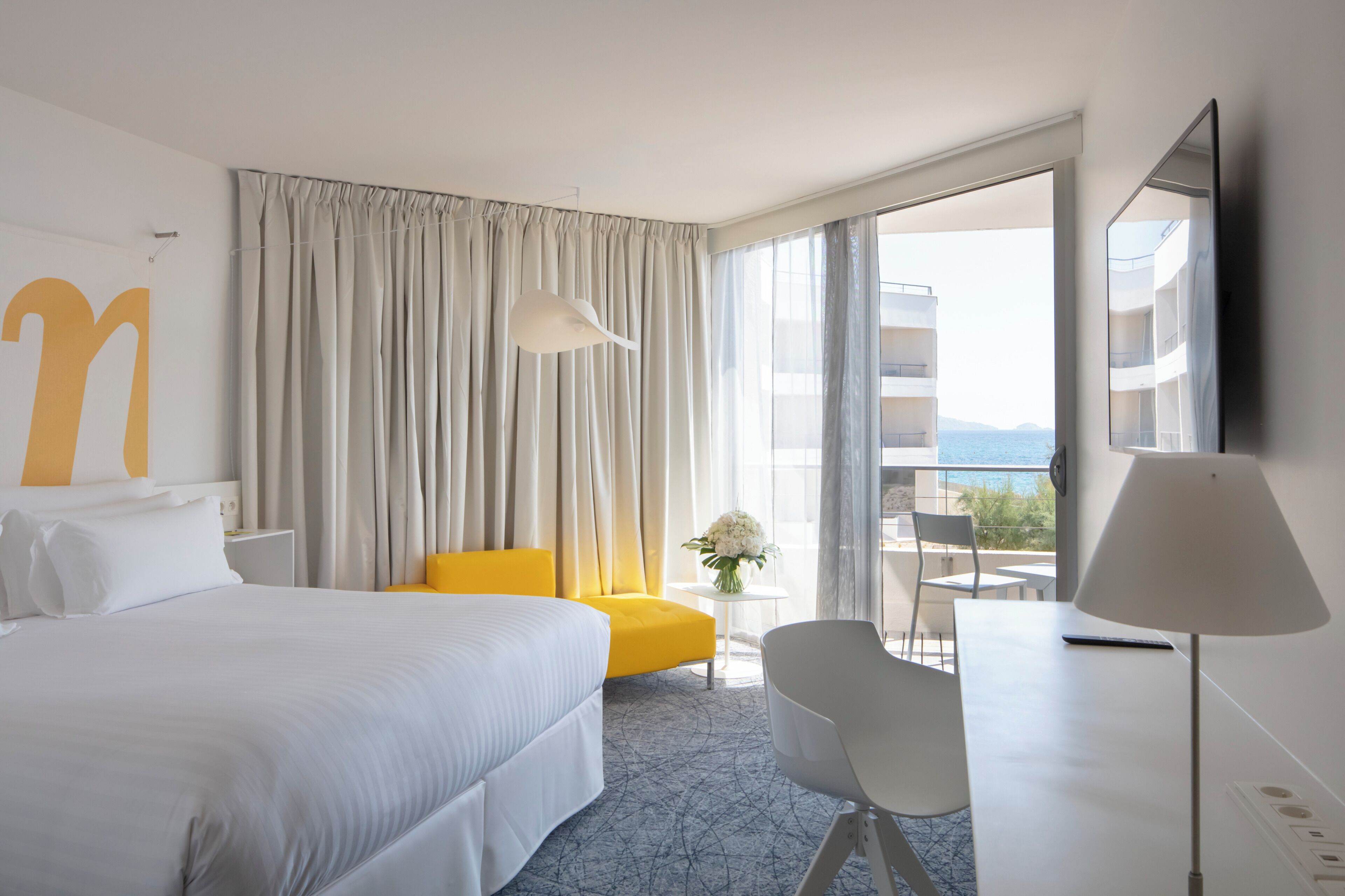 Una habitación de hotel con cama, escritorio, silla, lámpara, una pequeña mesa con un florero y un ventanal con cortinas.