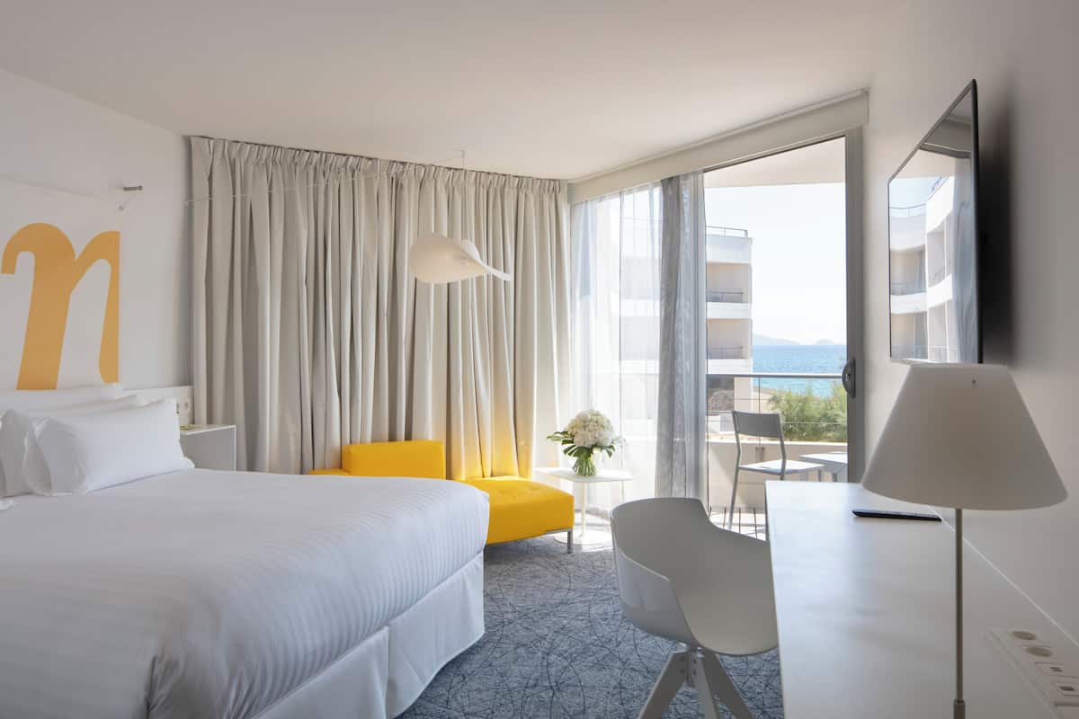 Una habitación de hotel con cama, escritorio, silla, lámpara, una pequeña mesa con un florero y un ventanal con cortinas.