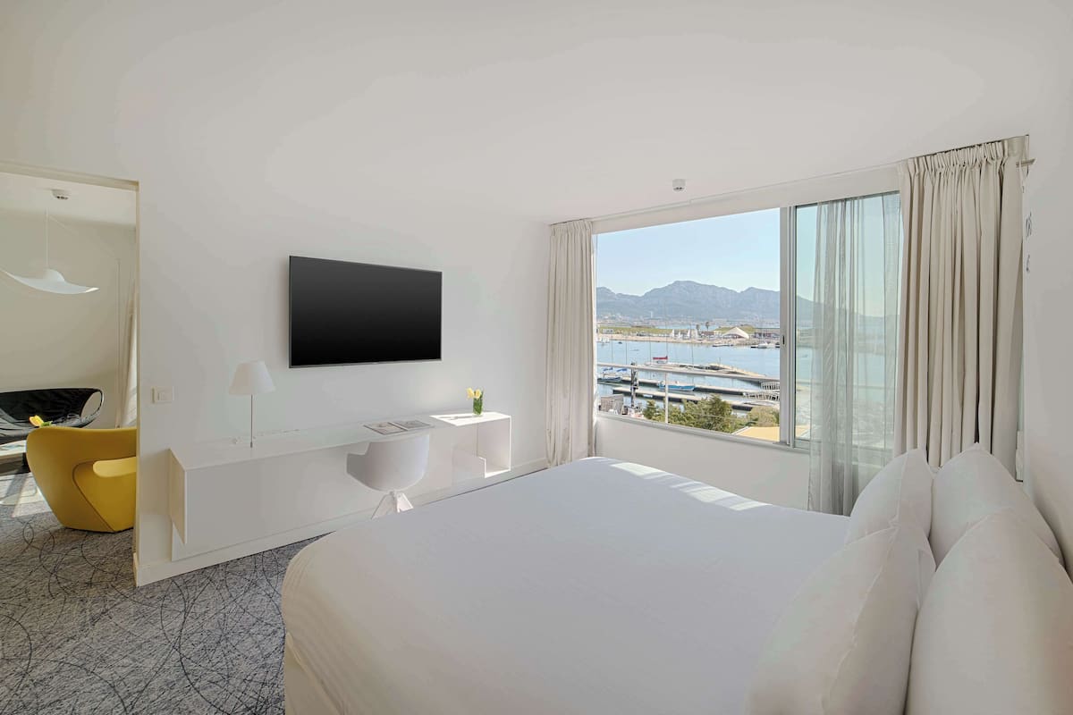 Habitación de hotel moderna con una cama grande, un televisor de pantalla plana en la pared, un escritorio pequeño blanco con una lámpara y vistas a un puerto deportivo y montañas a través de la ventana.