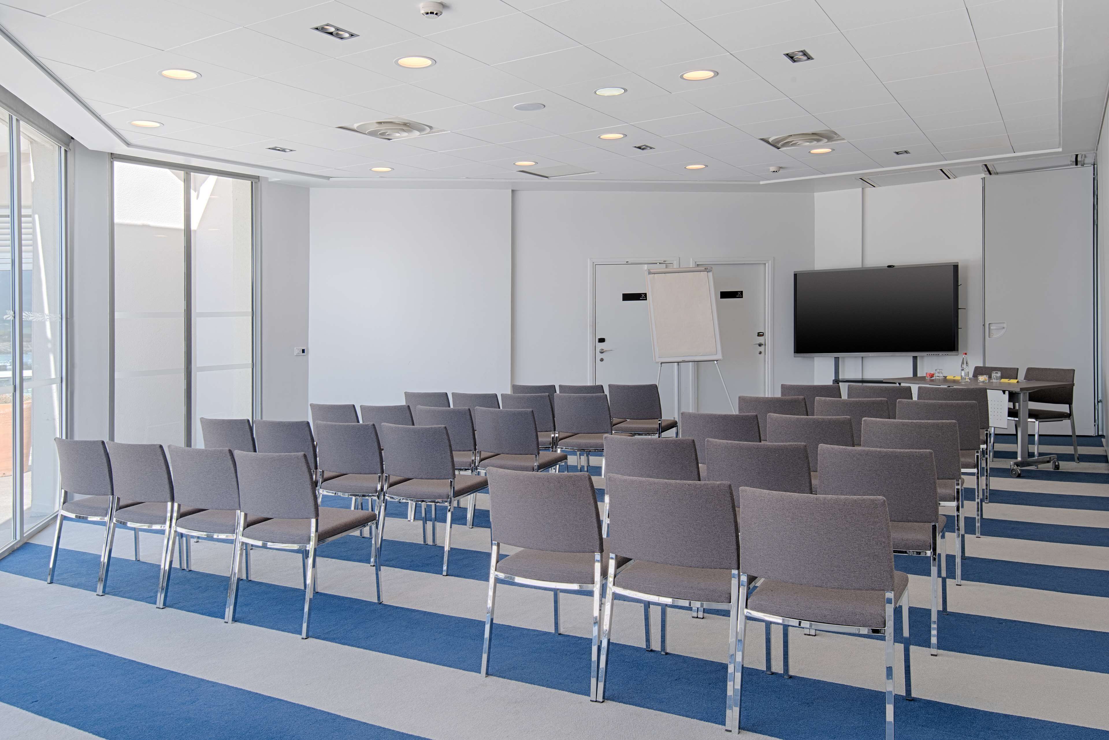 Une salle de conférence avec des rangées de chaises, un grand écran et un tableau blanc.