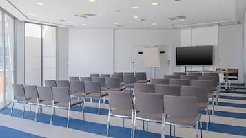 Une salle de conférence avec des rangées de chaises, un grand écran et un tableau blanc.