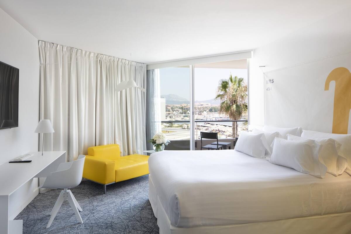 Habitación de hotel con una cama grande, un escritorio, una silla amarilla y vistas a la ciudad a través de ventanas amplias.