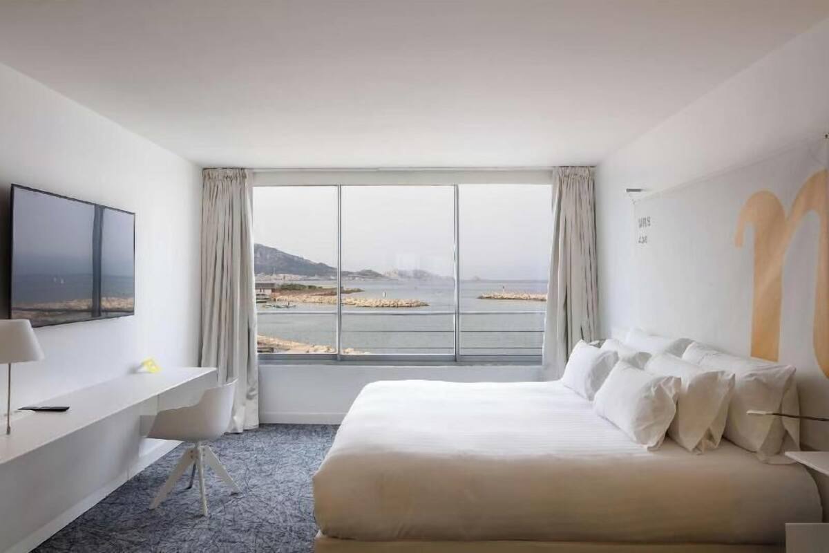 A hotel room with a large bed, a desk with a chair, a TV, and a view of the sea.