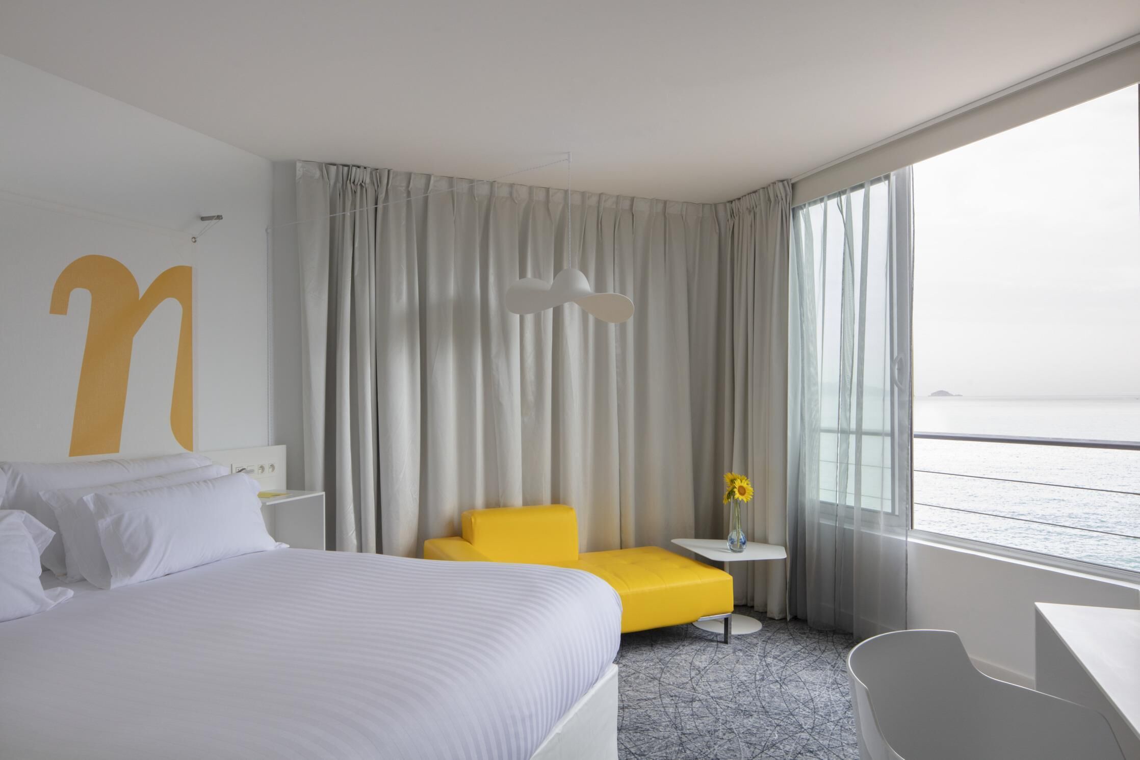 Habitación de hotel con una cama grande, un sillón amarillo, una mesita con un florero y vistas al mar.