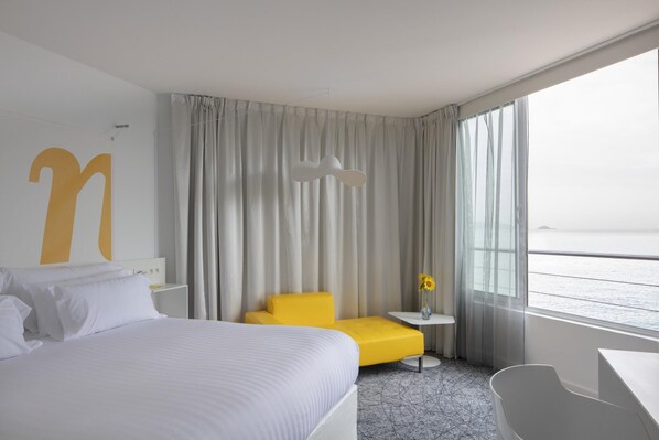 Habitación de hotel con una cama grande, un sillón amarillo, una mesita con un florero y vistas al mar.