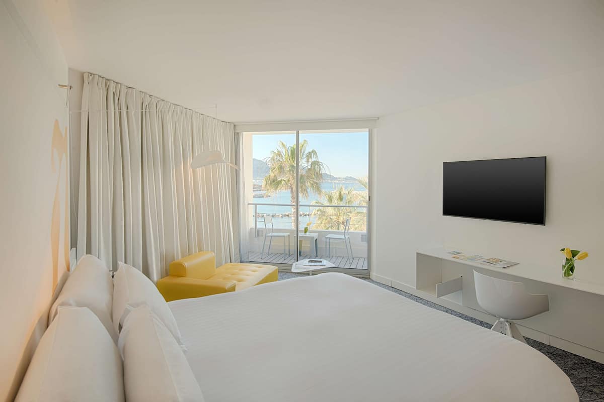 A hotel room with a large bed, a TV mounted on the wall, a balcony with a view of palm trees and mountains, and a small desk with a chair.