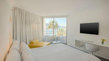 A hotel room with a large bed, a TV mounted on the wall, a balcony with a view of palm trees and mountains, and a small desk with a chair.