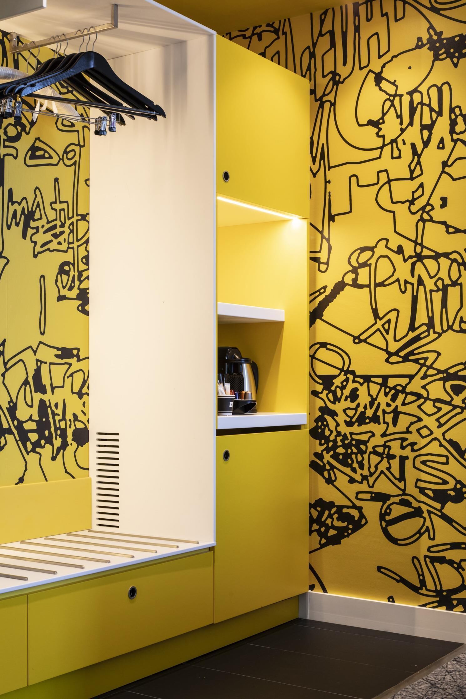 A room with a yellow wall featuring abstract black graffiti art, a white cabinet with a built-in heater, and a small shelf with a coffee maker.