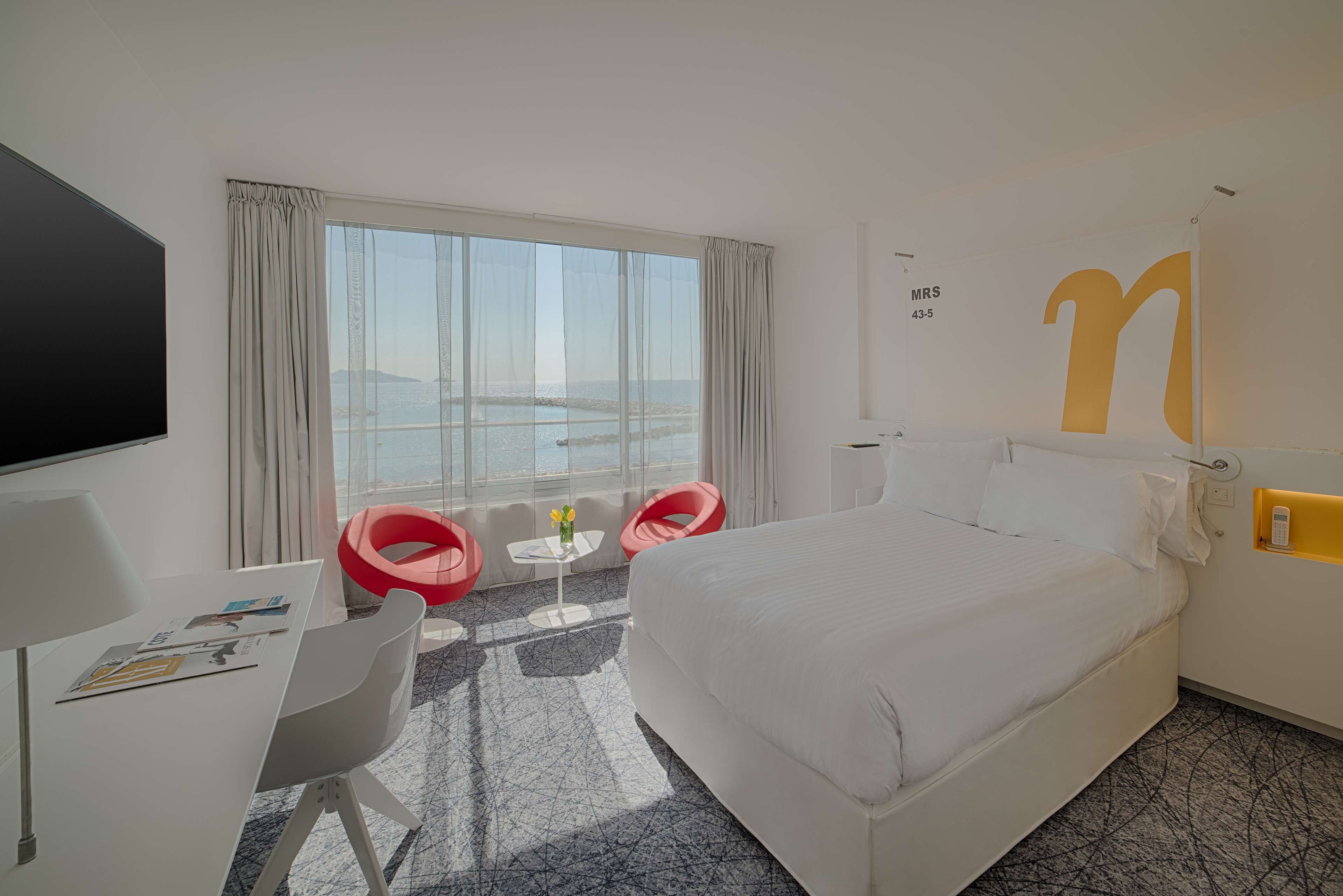 Habitación de hotel con una cama grande, un escritorio con televisor de pantalla plana, dos sillas rojas, una pequeña mesa con un florero y un ventanal con vistas al mar.