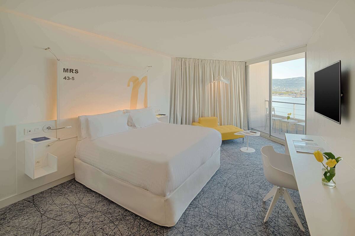 A hotel room with a large bed, a desk, a chair, a TV, and a window with a view of the sea.