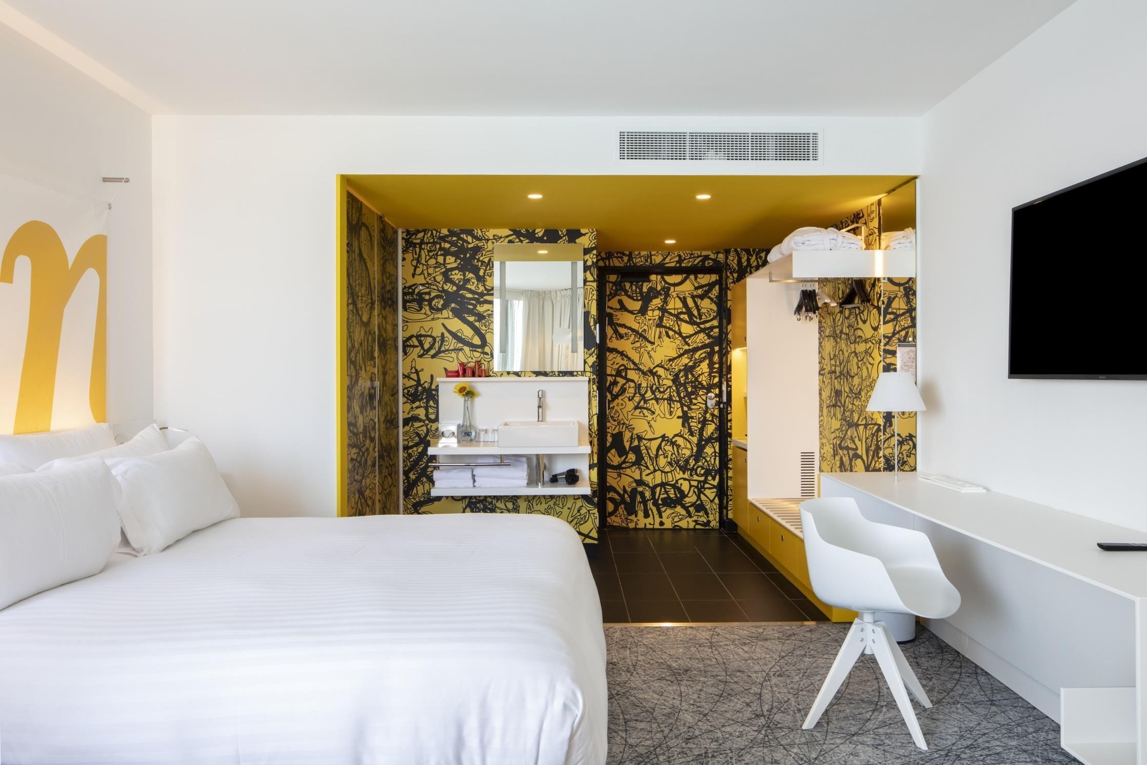A modern hotel room with a large bed, a desk, and a bathroom with a mirror and sink.