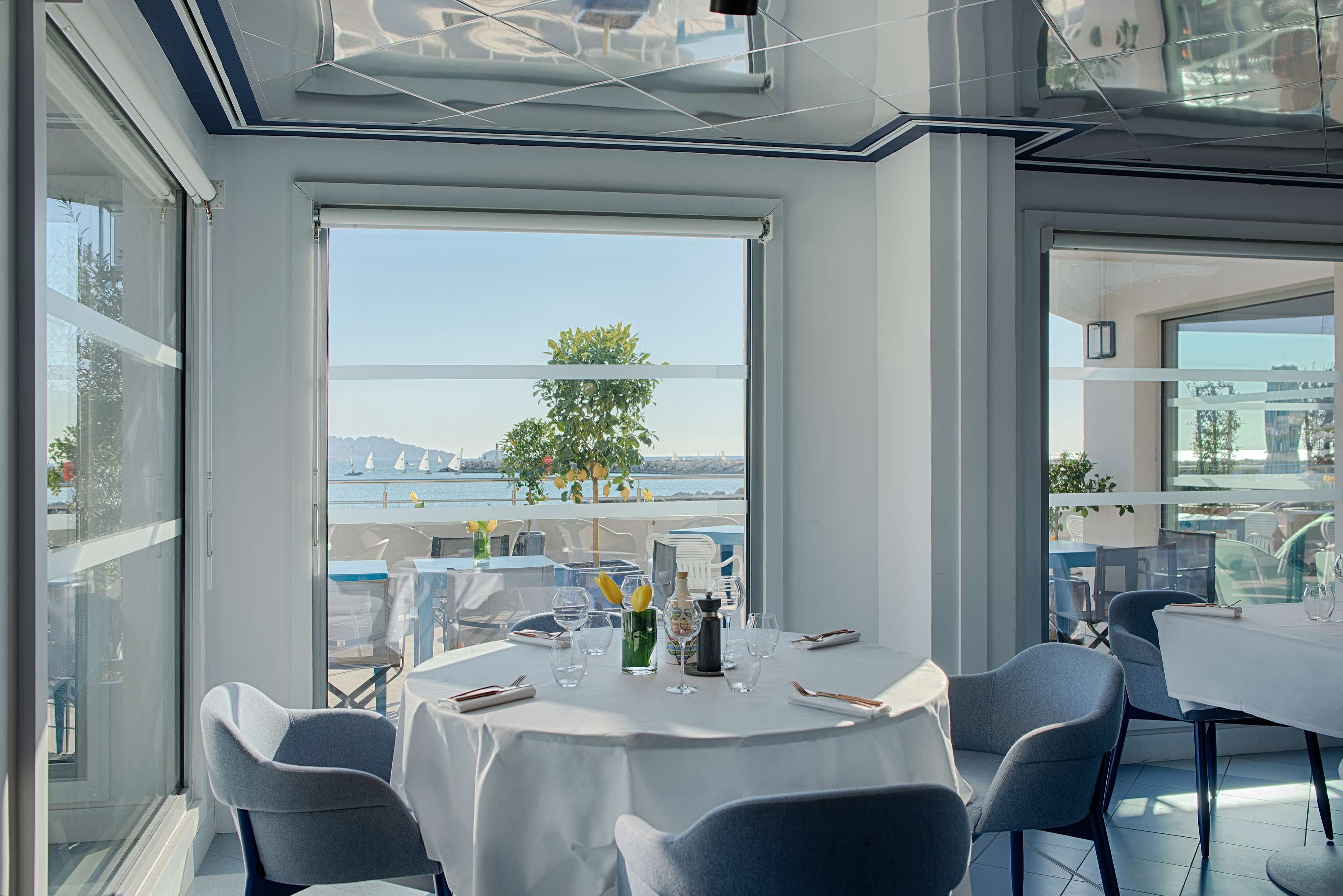 A dining area with tables set for two, large windows offering a view of the sea and hills, and a modern interior design.