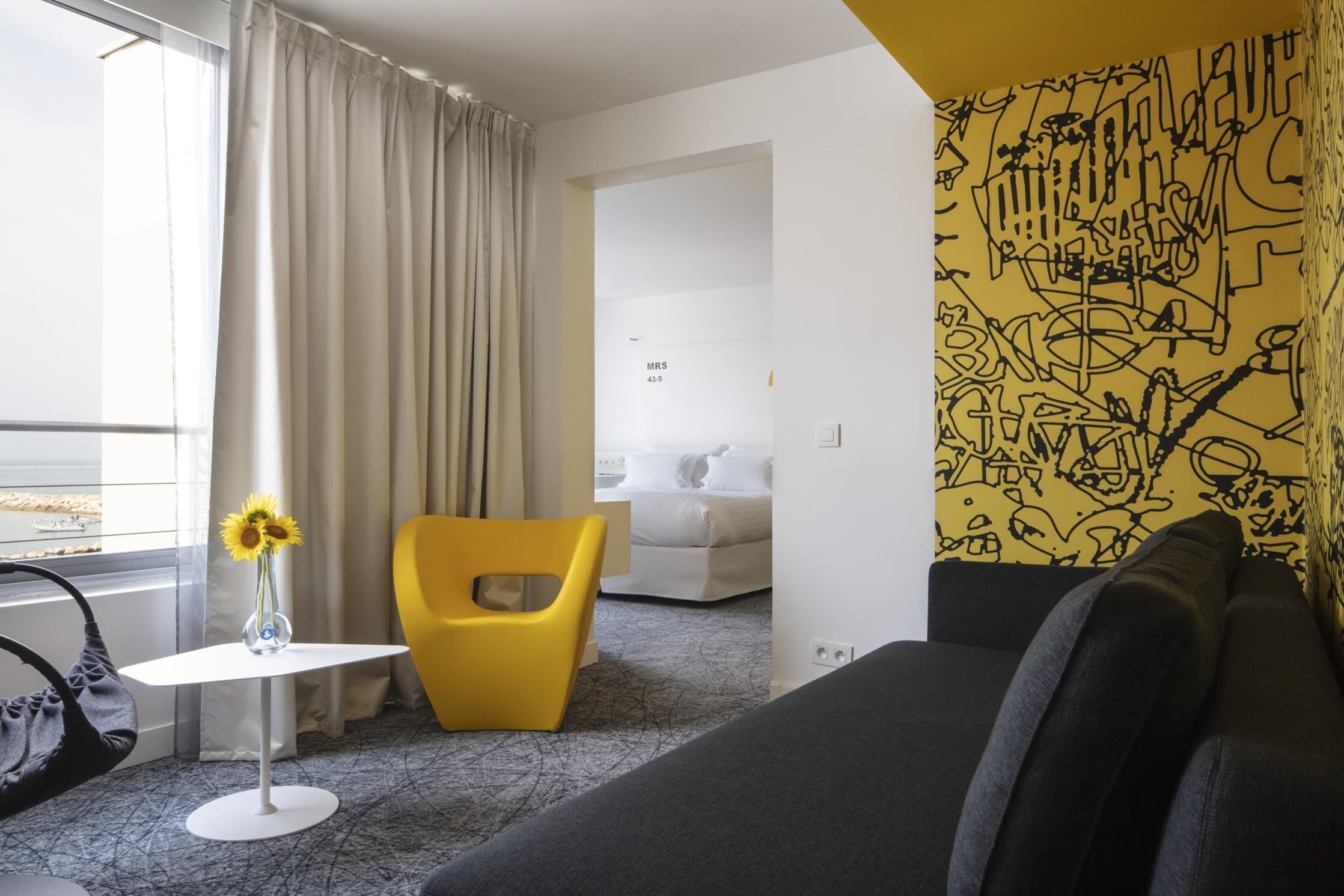 A hotel room with a modern design, featuring a black sofa, a yellow chair, a small white table with a vase of sunflowers, and a large window with curtains.