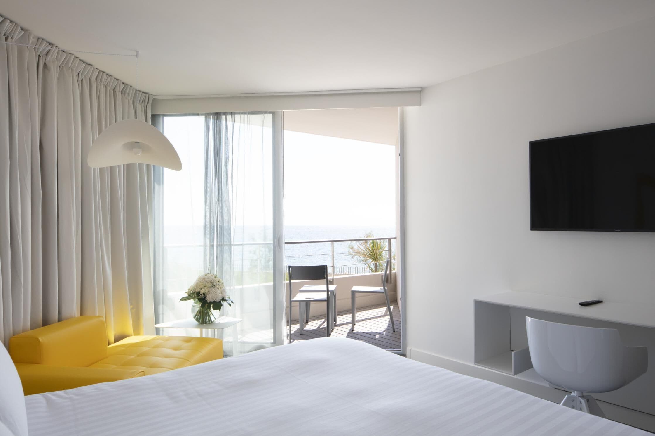 Habitación de hotel con cama, un sillón amarillo, una silla blanca, una mesita con un florero y un balcón con vista al mar.