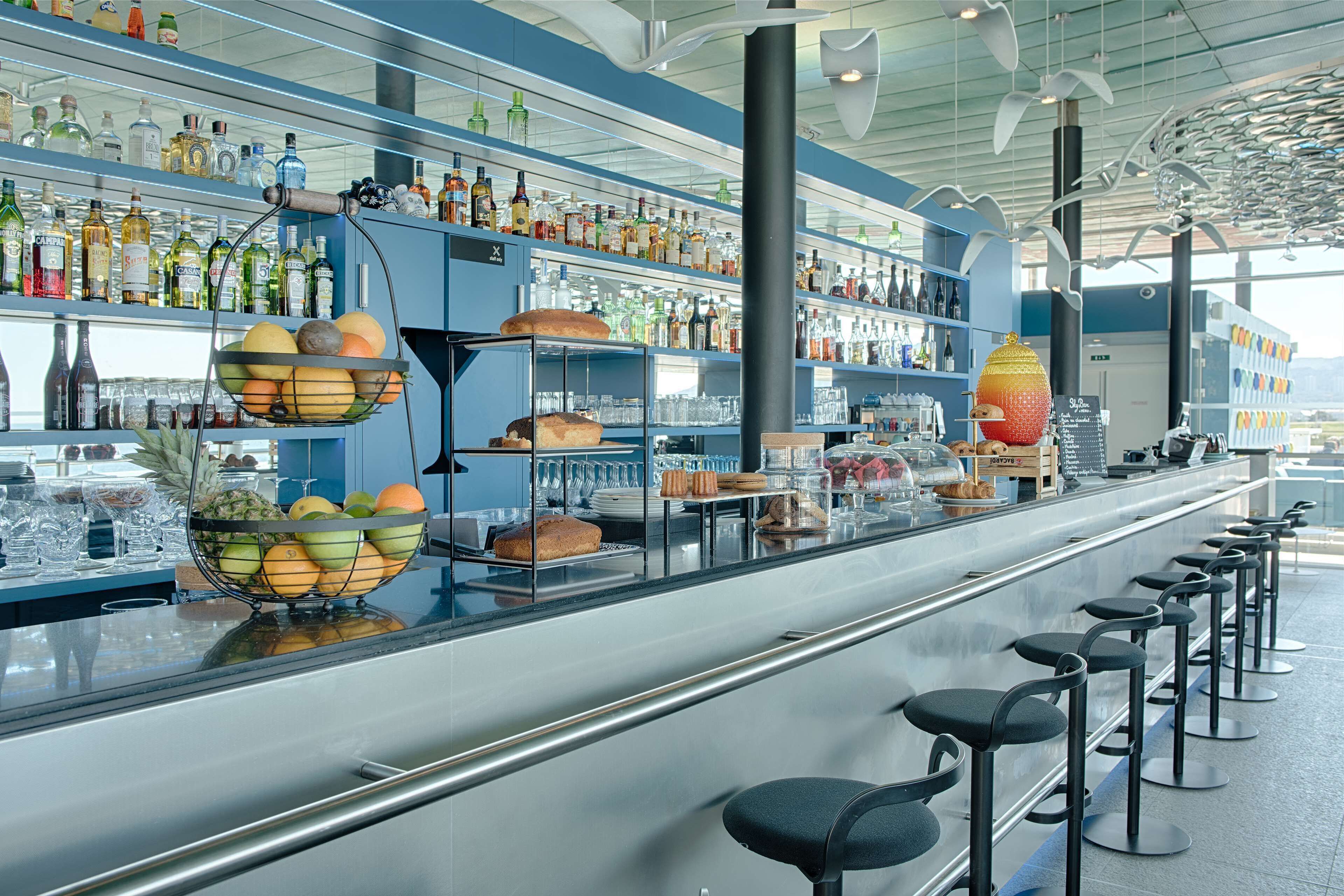 Un espace bar doté d’un long comptoir, de nombreux tabourets et d’étagères remplies de diverses bouteilles et de fruits.