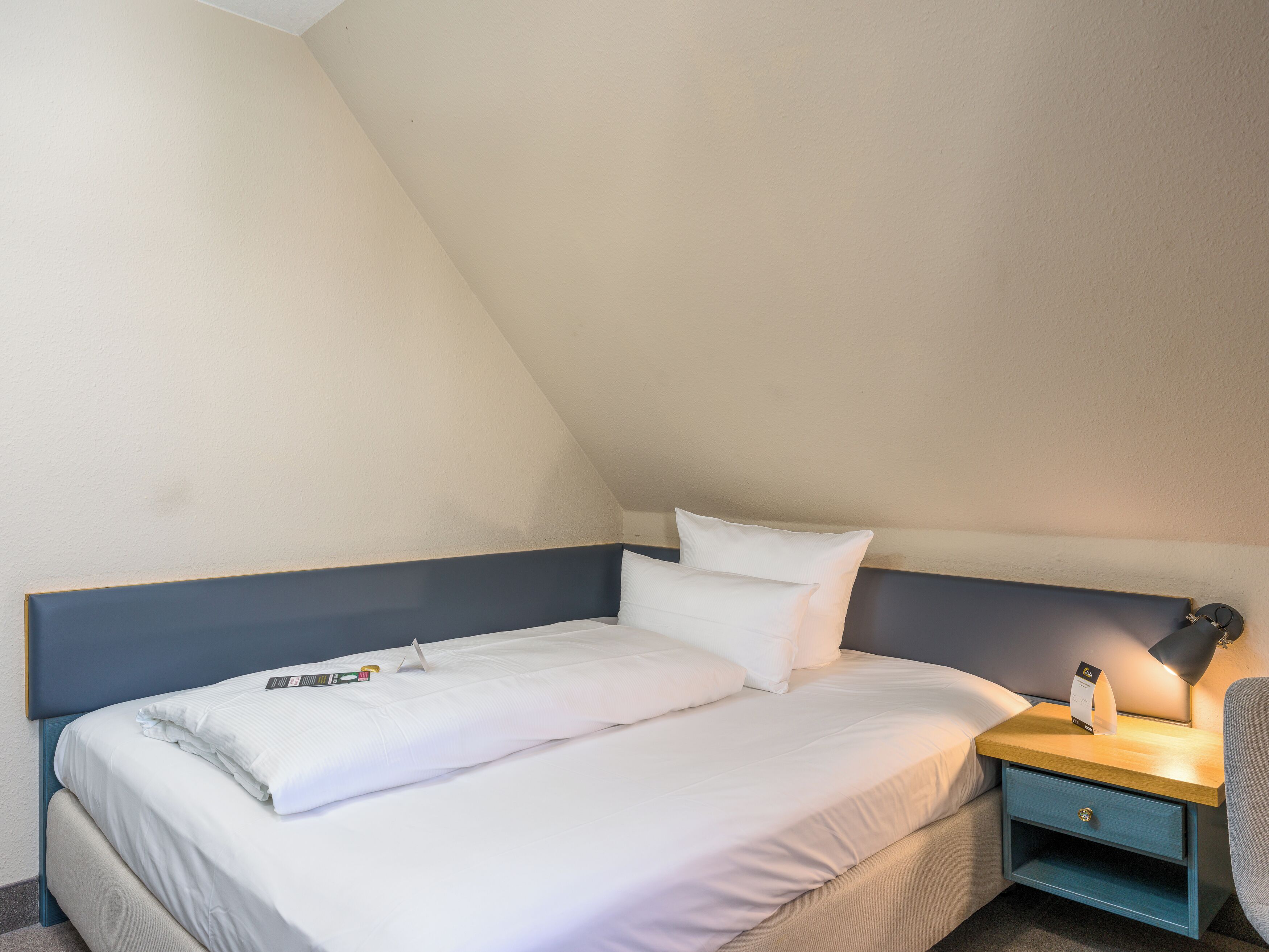 Een net opgemaakt bed met witte lakens, een nachtkastje met een lamp en een klein ladeblok.
