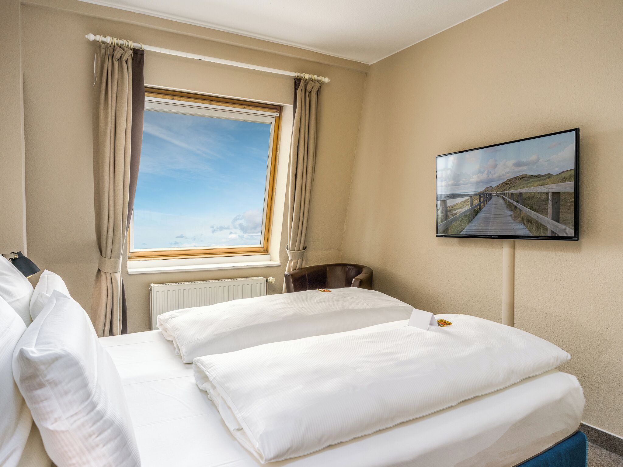 Et hotelværelse med en seng, et vindue med gardiner, en radiator, en stol og et billede i ramme på væggen.