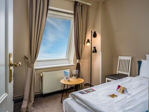 A hotel room with a bed, a small table, a chair, a radiator, and a window with curtains. - PLAZA Premium Sylt (Wenningstedt-Braderup)