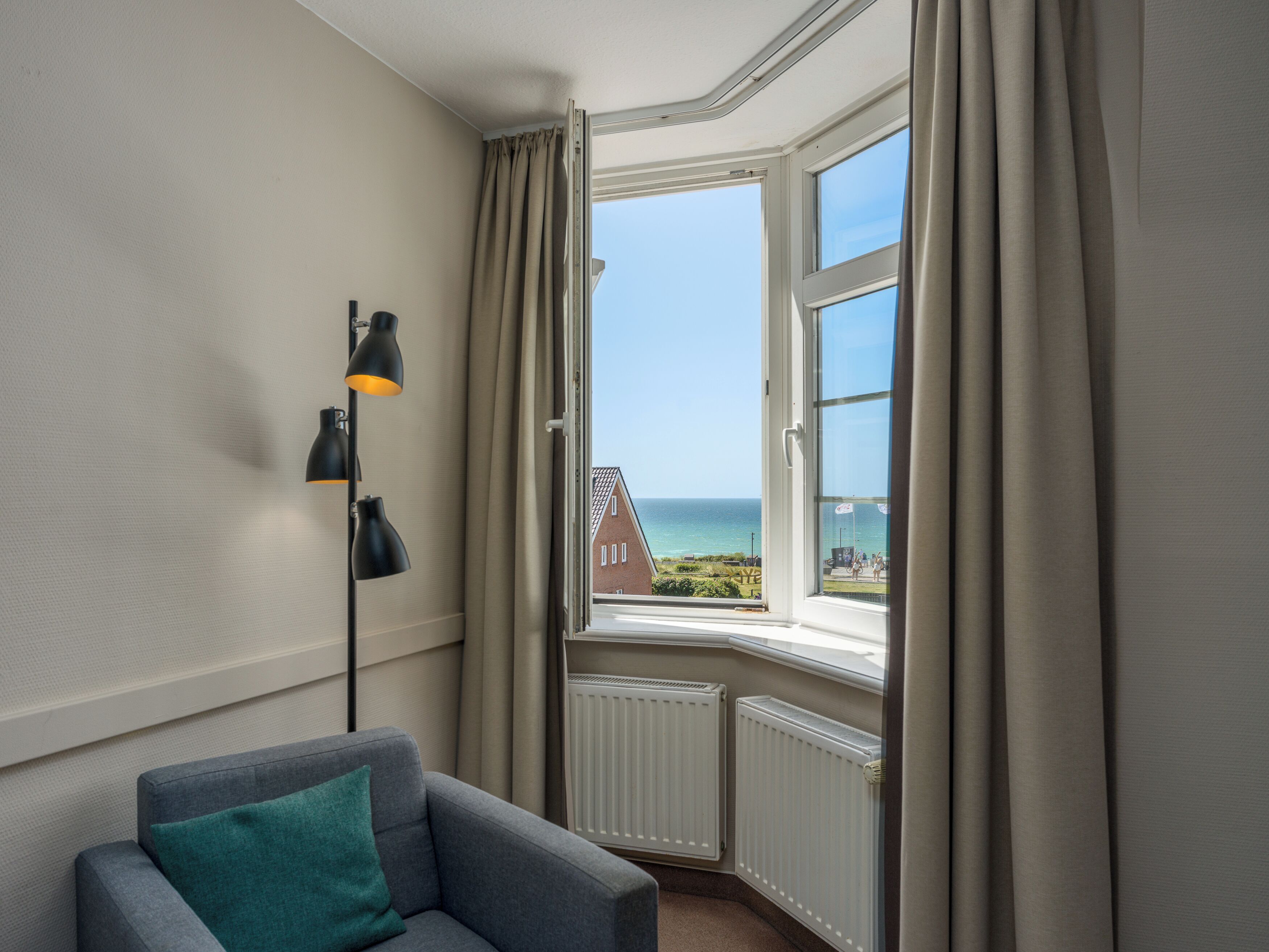 A room with a corner window, a grey sofa with a teal pillow, and a black floor lamp.