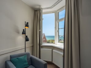 A room with a corner window, a grey sofa with a teal pillow, and a black floor lamp. - PLAZA Premium Sylt (Wenningstedt-Braderup)