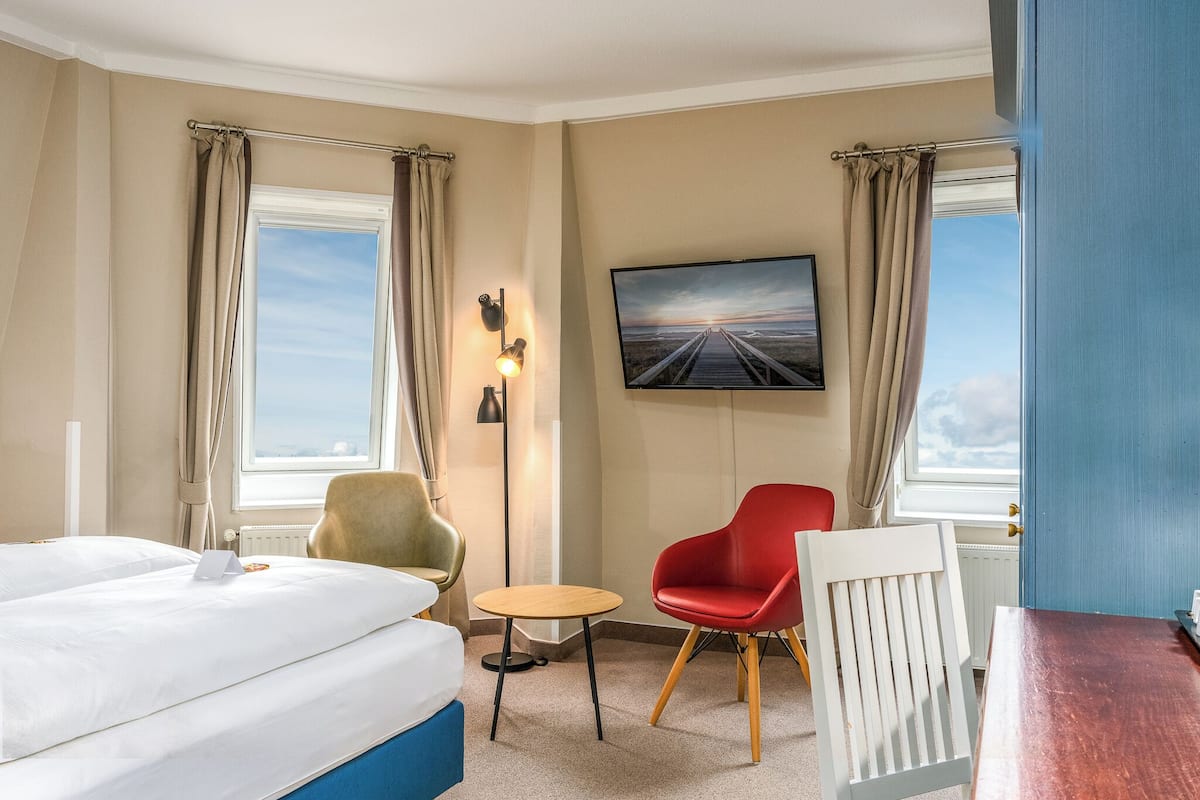 A hotel room with a bed, a desk, a chair, a small table, a TV mounted on the wall, and a view of the beach through the windows.
