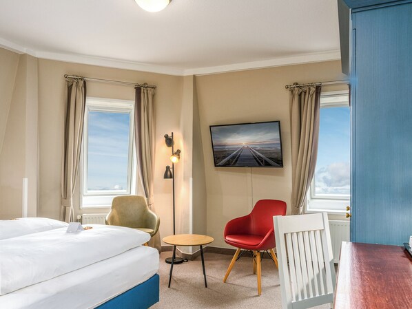 A hotel room with a bed, a desk, a chair, a small table, a TV mounted on the wall, and a view of the beach through the windows. - PLAZA Premium Sylt (Wenningstedt-Braderup)