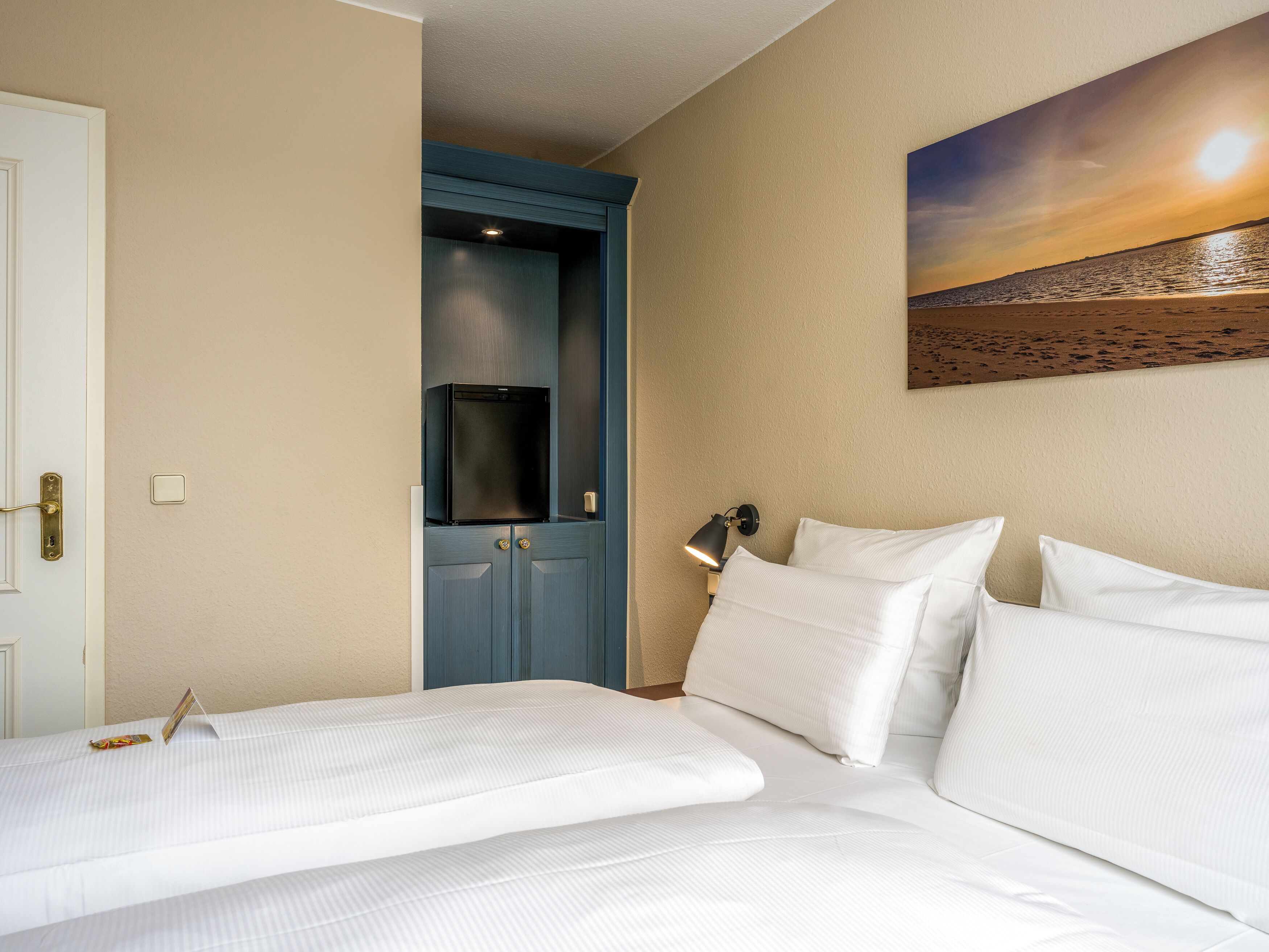 A hotel room with a bed, a door, a wall-mounted TV, a nightstand with a lamp, and a painting of a sunset.