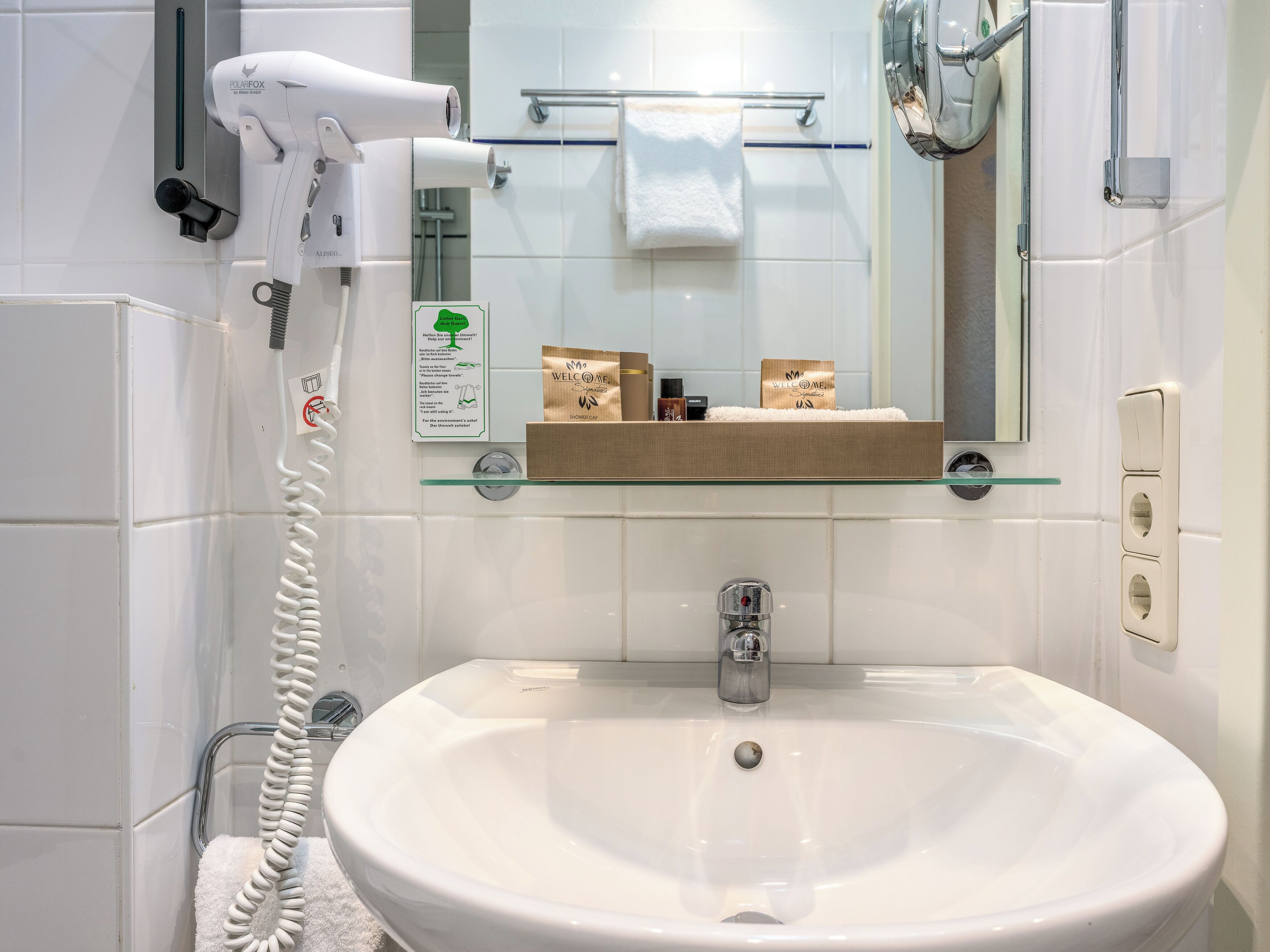 A bathroom with a white sink, a glass shelf, a towel rack, a hairdryer, and a shower area.