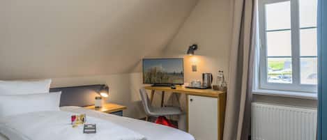 Quarto de hotel com cama, escrivaninha com computador, cadeira, janela com cortinas e radiador.