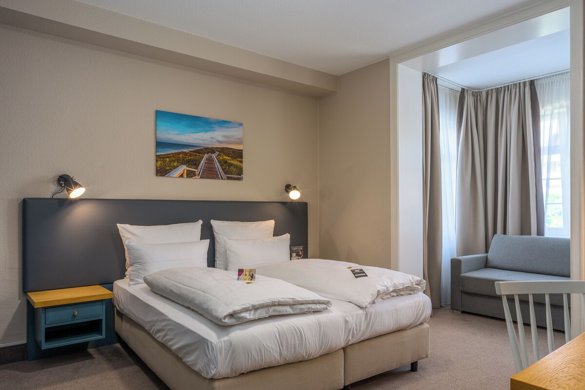 A hotel room with a bed, bedside tables, a desk, a chair, and a painting on the wall.