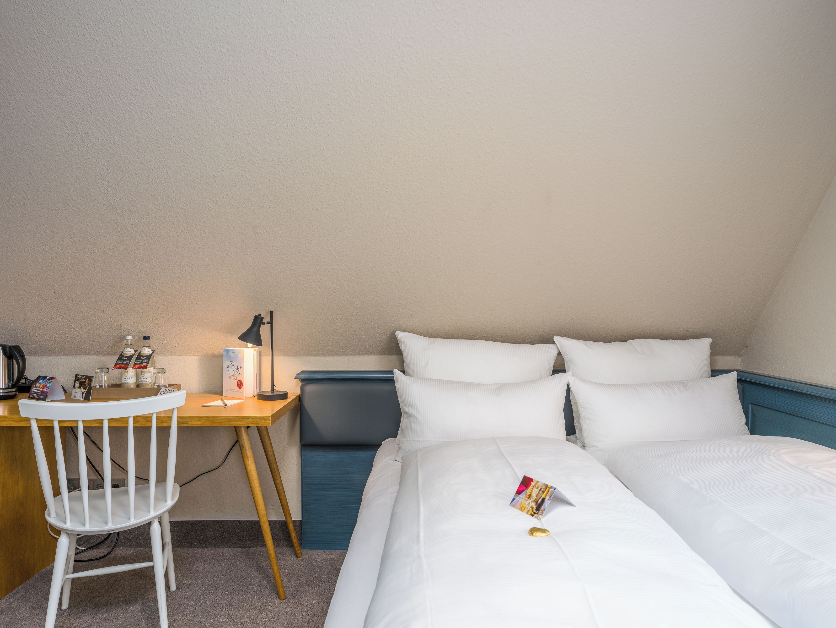 A hotel room with a bed, a desk, a chair, a lamp, and a bedside table with a book and a bottle.