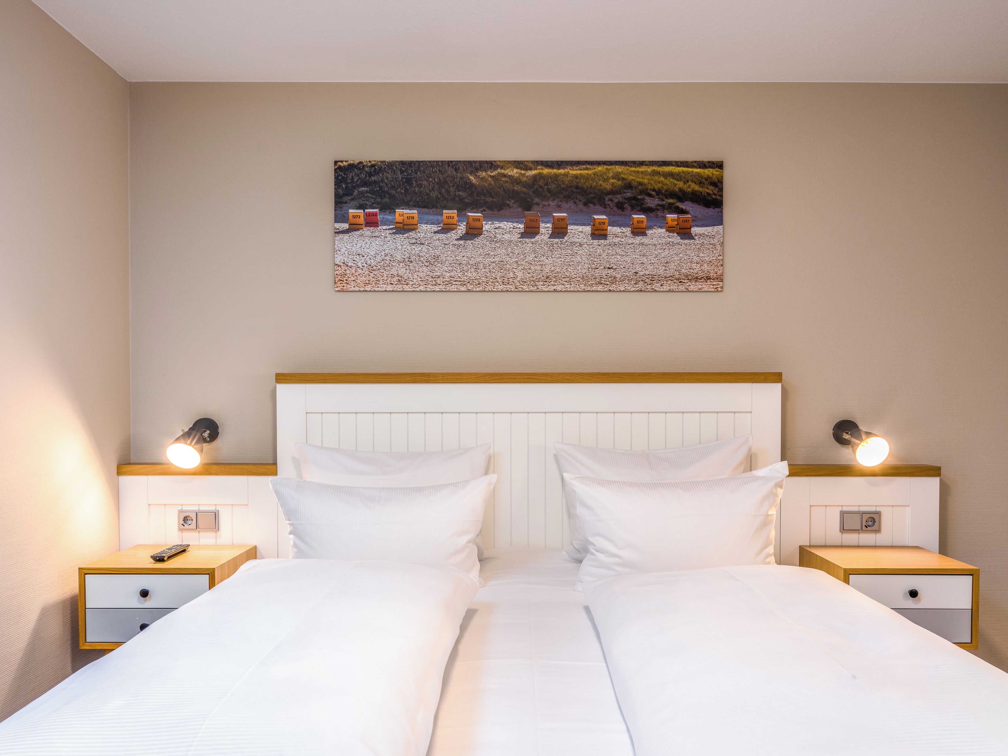 Un lit double avec du linge de lit blanc, deux tables de chevet avec des lampes, une tête de lit et un tableau représentant des cabanes de plage accroché au mur.