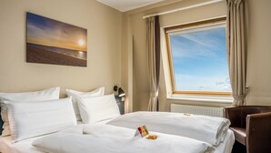 A hotel room with a bed, a desk, a chair, a window with curtains, and a painting on the wall.