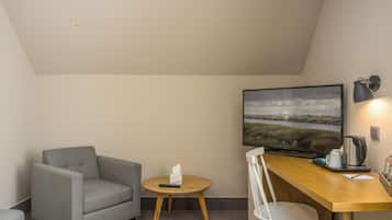 Ein Zimmer mit grauem Sessel, einem kleinen runden Tisch mit Fernbedienung und Taschentüchern, einem Holztisch mit Stuhl, einem Flachbildfernseher, der eine Landschaft zeigt, und einer Wandlampe.