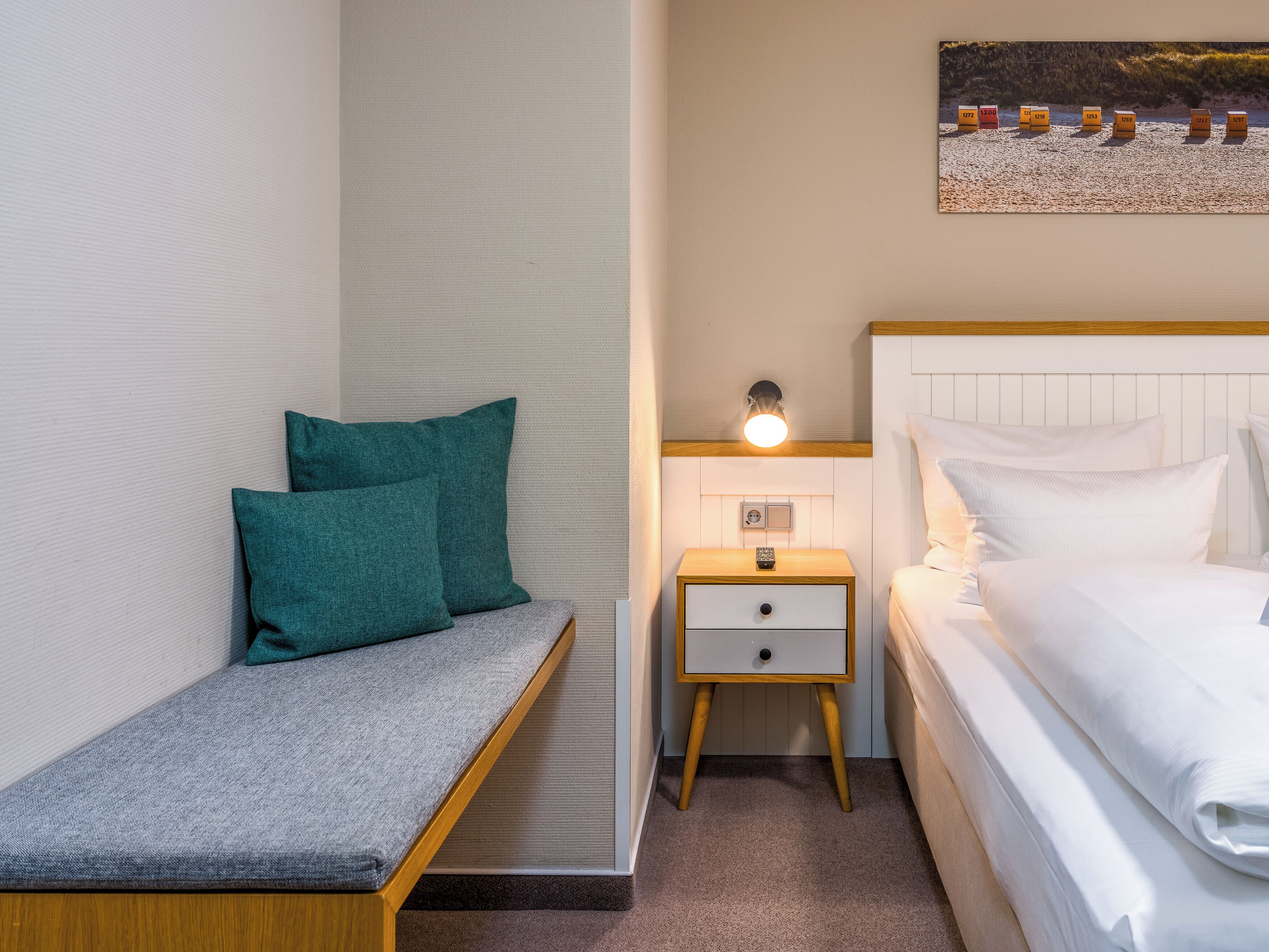 A bedroom with a bed, a bedside table with a lamp, a nightstand, a bench with pillows, and a wall-mounted artwork.