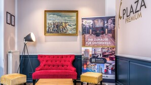 A hotel lobby with a red sofa, a standing lamp, and a poster advertising the hotel.
