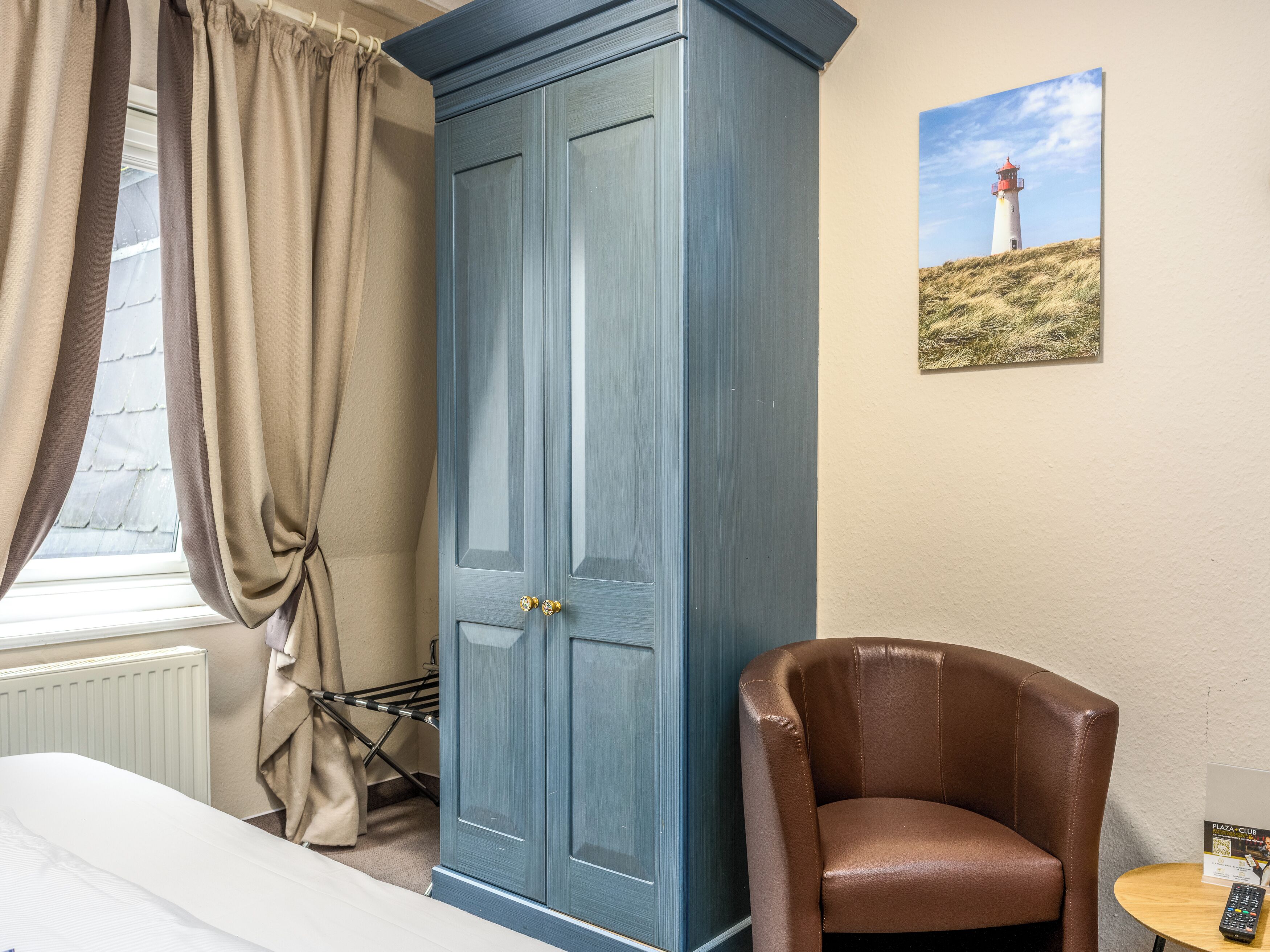 A room with a large blue wardrobe, a brown leather chair, a small round table with a remote control, and a painting of a lighthouse on the wall.