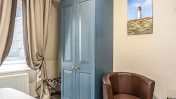 A room with a large blue wardrobe, a brown leather chair, a small round table with a remote control, and a painting of a lighthouse on the wall.
