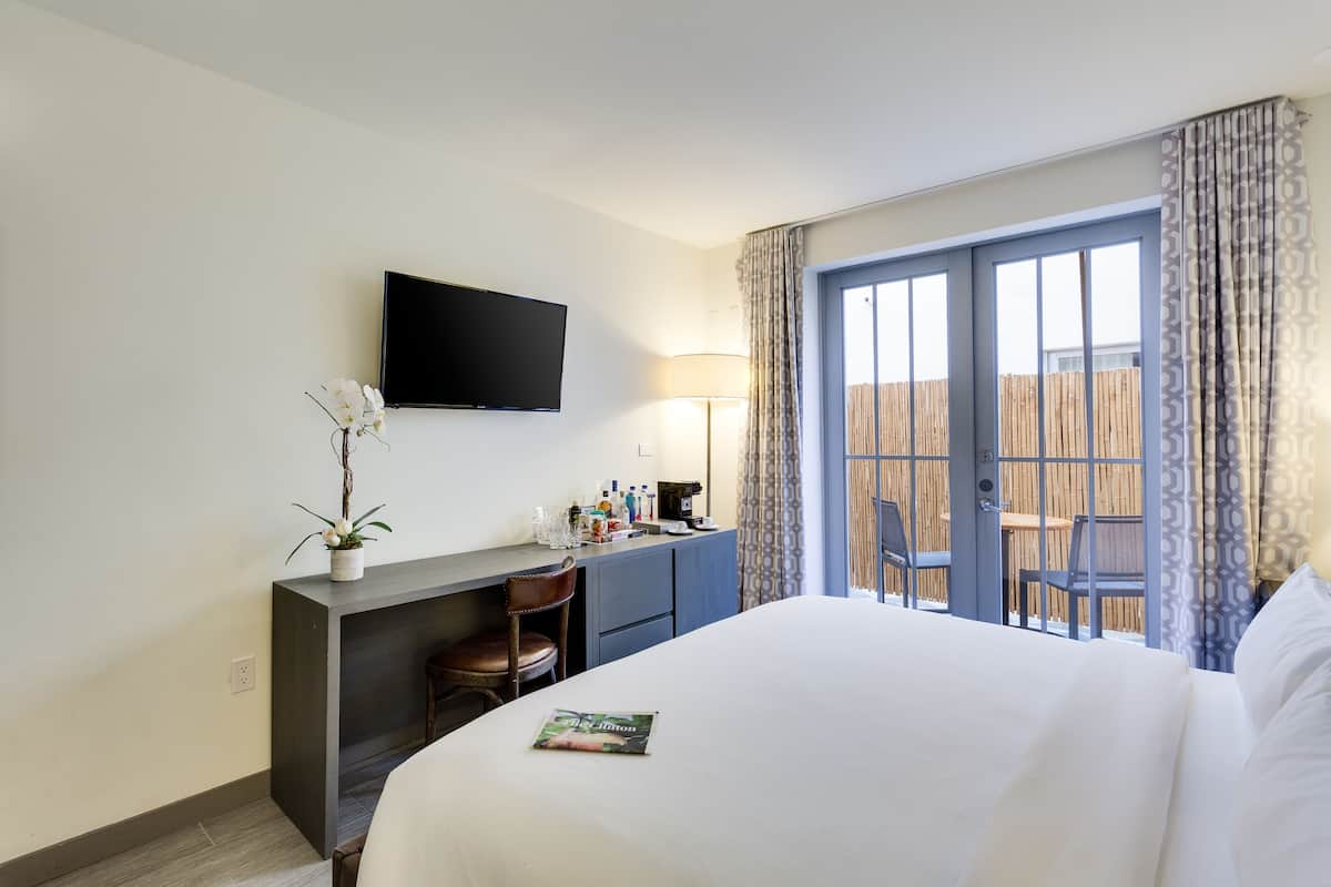 Habitación de hotel con una cama, un escritorio con silla, un televisor montado en la pared y un balcón con vista a una cerca de madera.