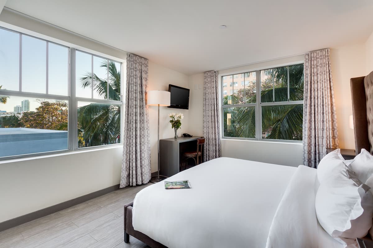 Habitación de hotel con una cama grande, un escritorio con lámpara, televisor y vistas a palmeras y al paisaje urbano.
