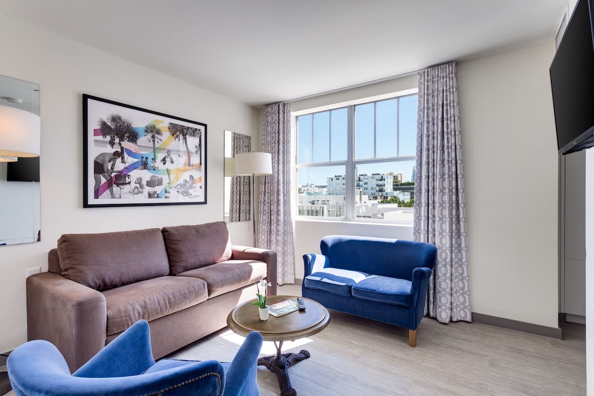 Una sala de estar moderna con un sofá marrón, un sillón azul y una mesita de centro. Un ventanal ofrece vistas a edificios.