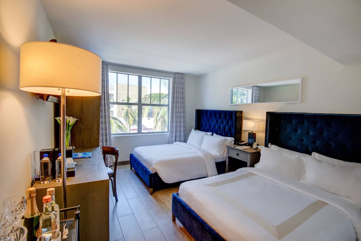 Habitación de hotel con dos camas, un escritorio con lámpara, un minibar y un ventanal con vistas a palmeras.