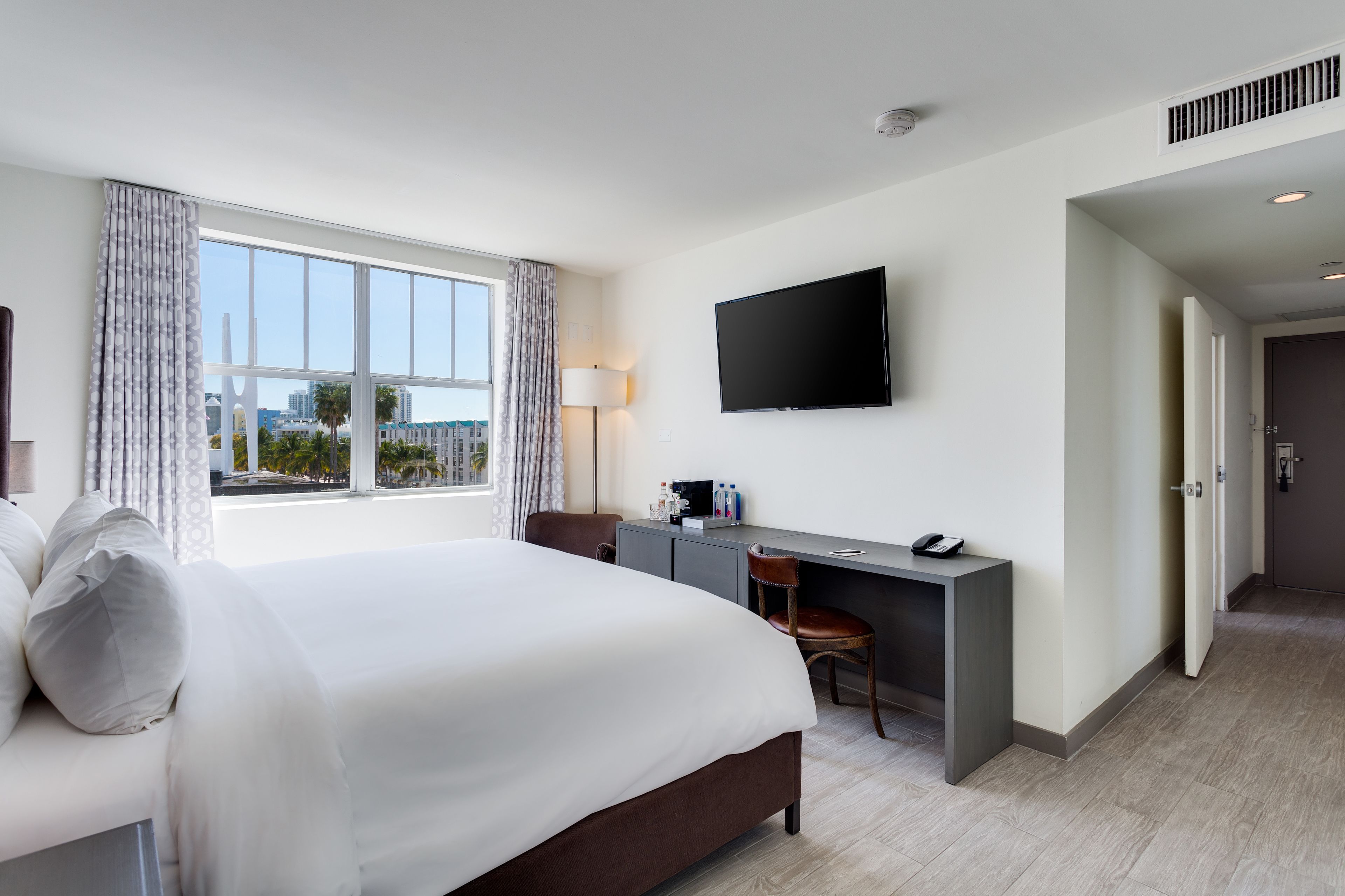 A modern hotel room with a large bed, a desk, a chair, a flat-screen TV, and a view of the city.