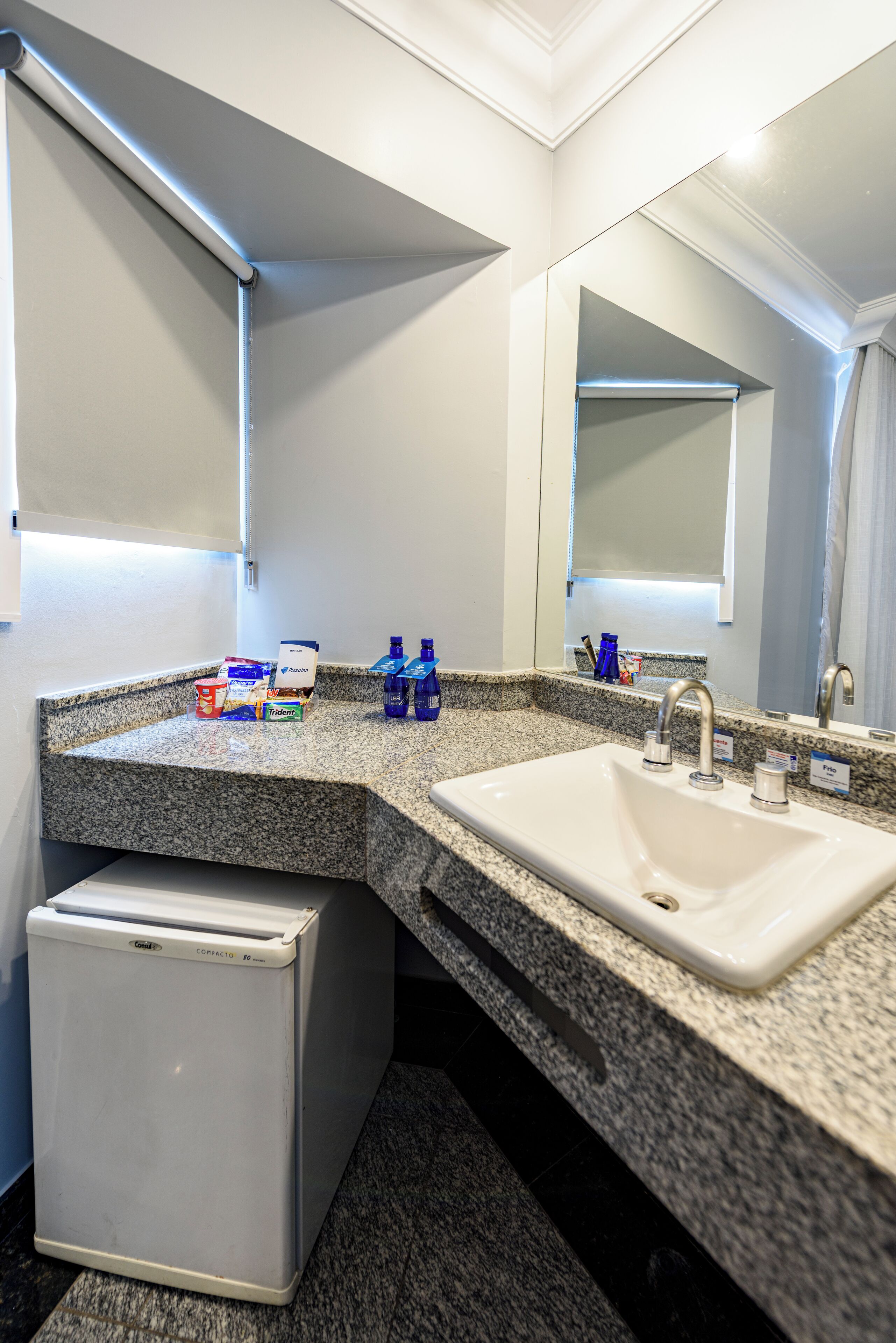 A bathroom with a granite countertop, two sinks, a mirror, and a compact refrigerator.