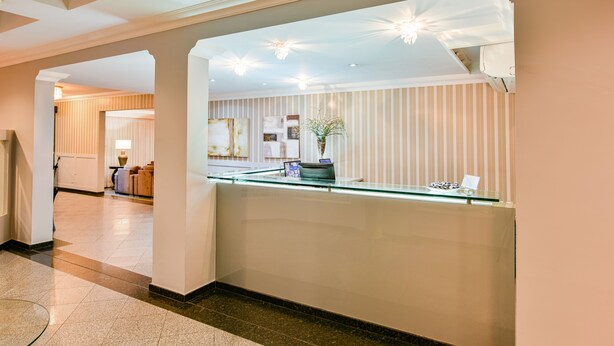 Zona de recepción de hotel con un separador de vidrio, mostrador de recepción y una planta decorativa.