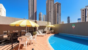 Zona de piscina en la azotea con sillones y sombrillas, rodeada de edificios altos.