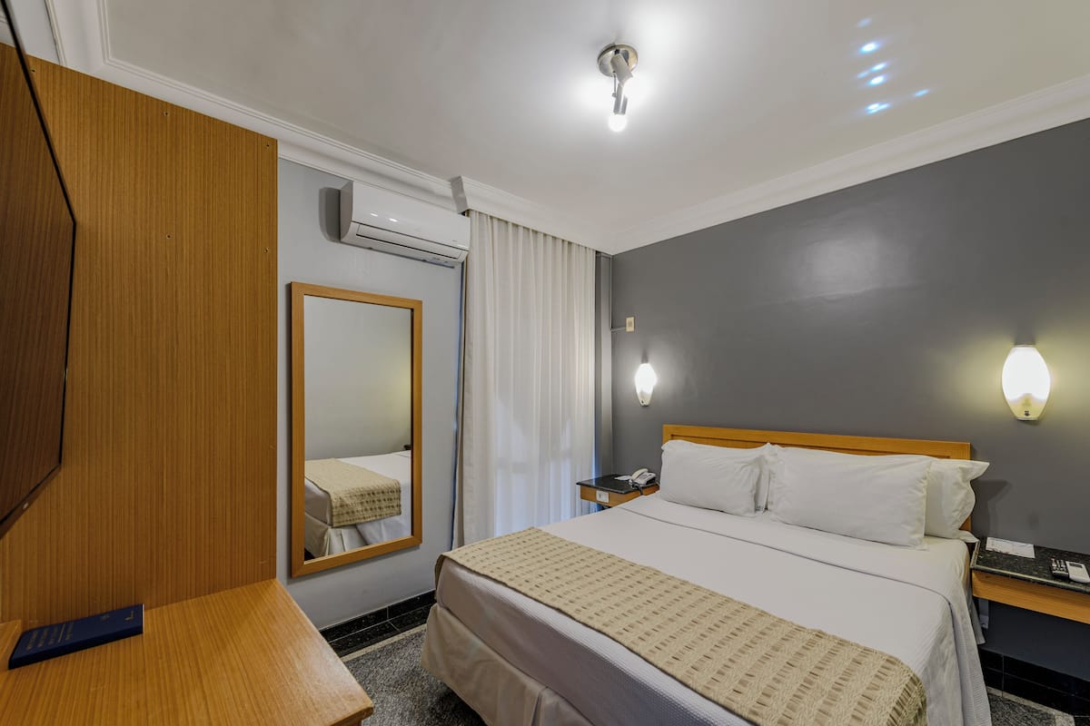 Ein Hotelzimmer mit einem großen Bett, einem Holzschrank, einem Nachttisch mit Lampe, einem Spiegel und einer Wand-Klimaanlage.