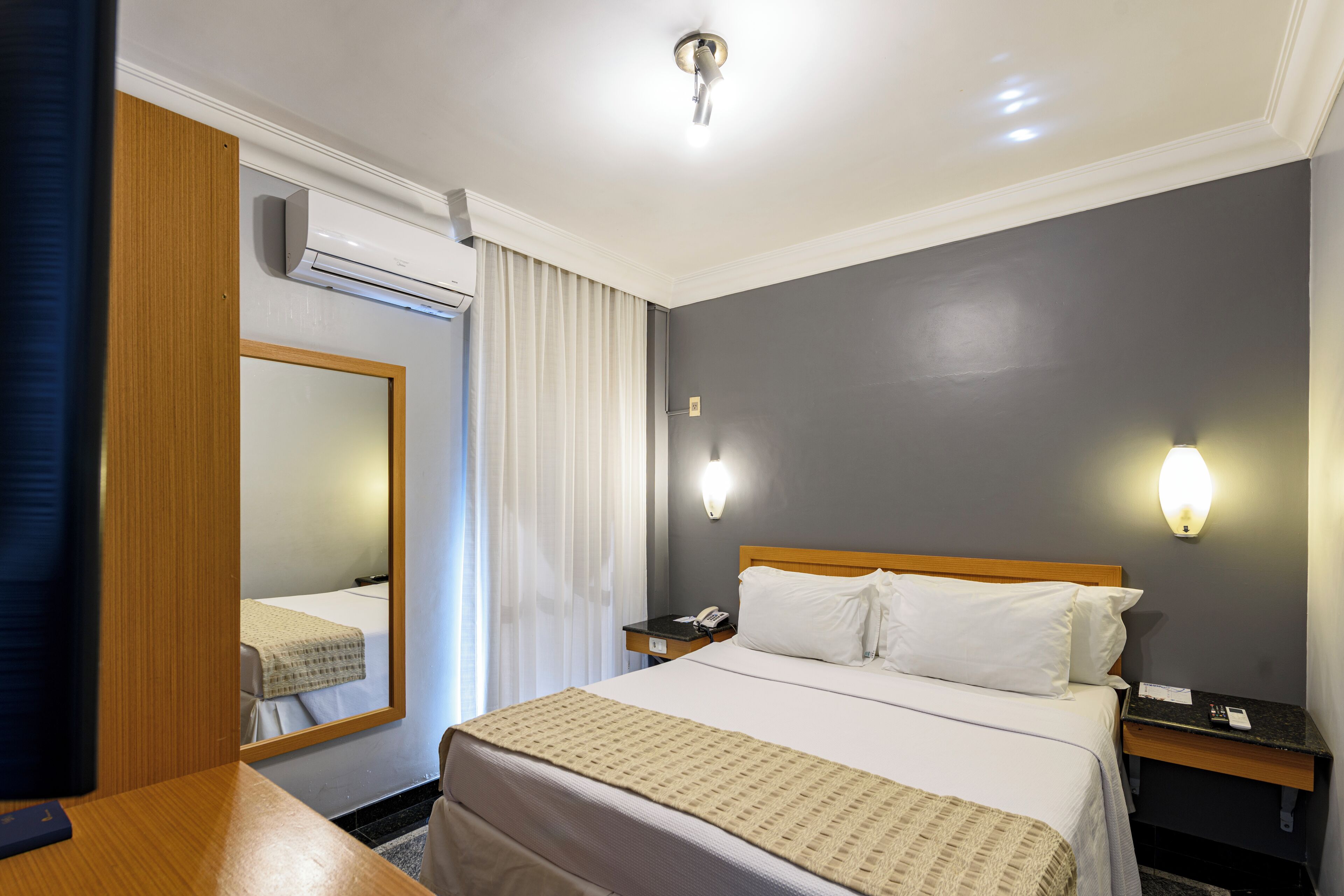 Habitación de hotel con cama, mesitas de noche, espejo, equipo de aire acondicionado y lámpara de pared.
