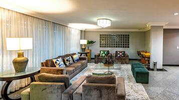 A modern living room with a sectional sofa, a round coffee table, and a green ottoman. There is a dining area with a wooden table and chairs in the background.