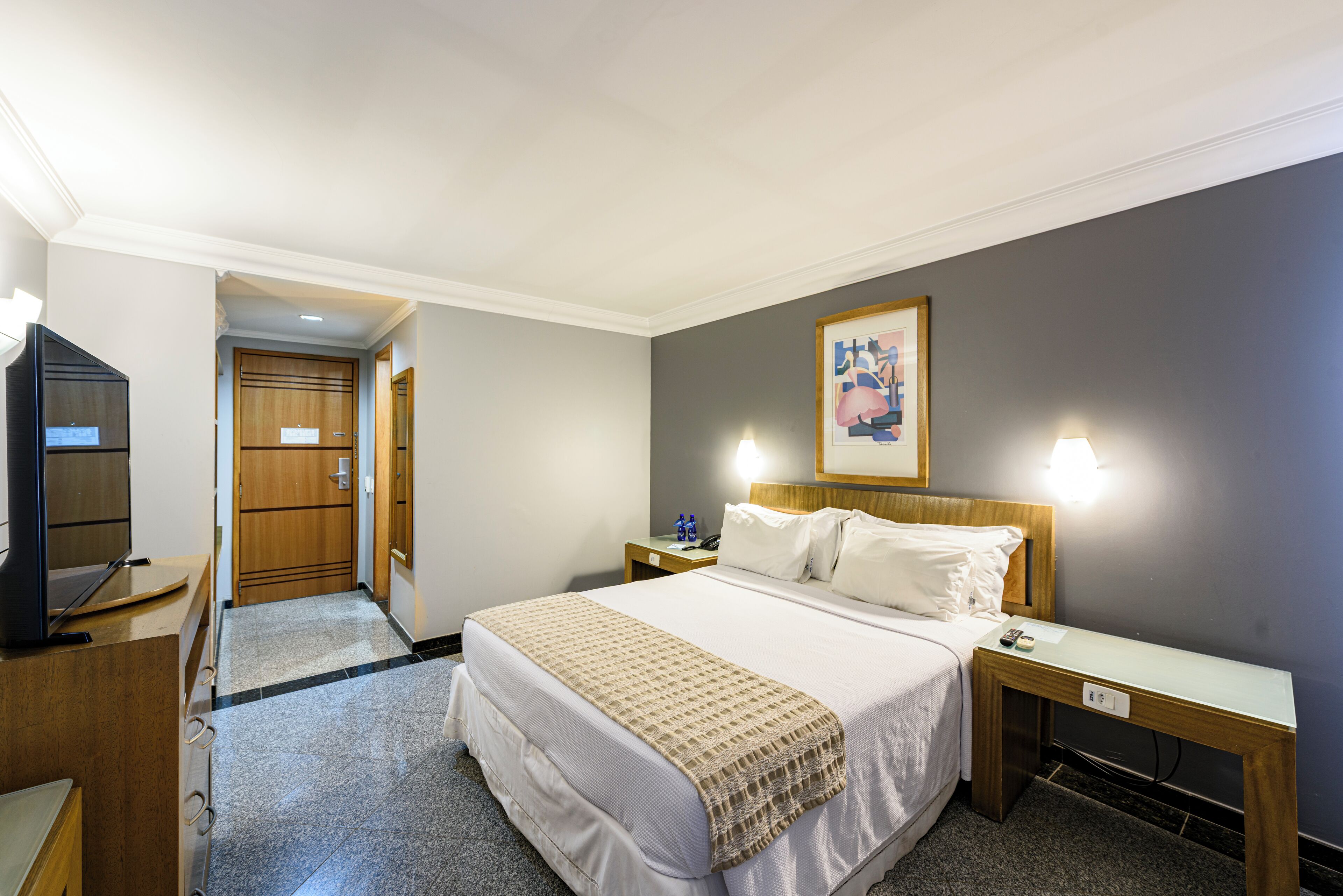 Une chambre d’hôtel avec un lit, des tables de chevet, un bureau, une télévision et un tableau au mur.